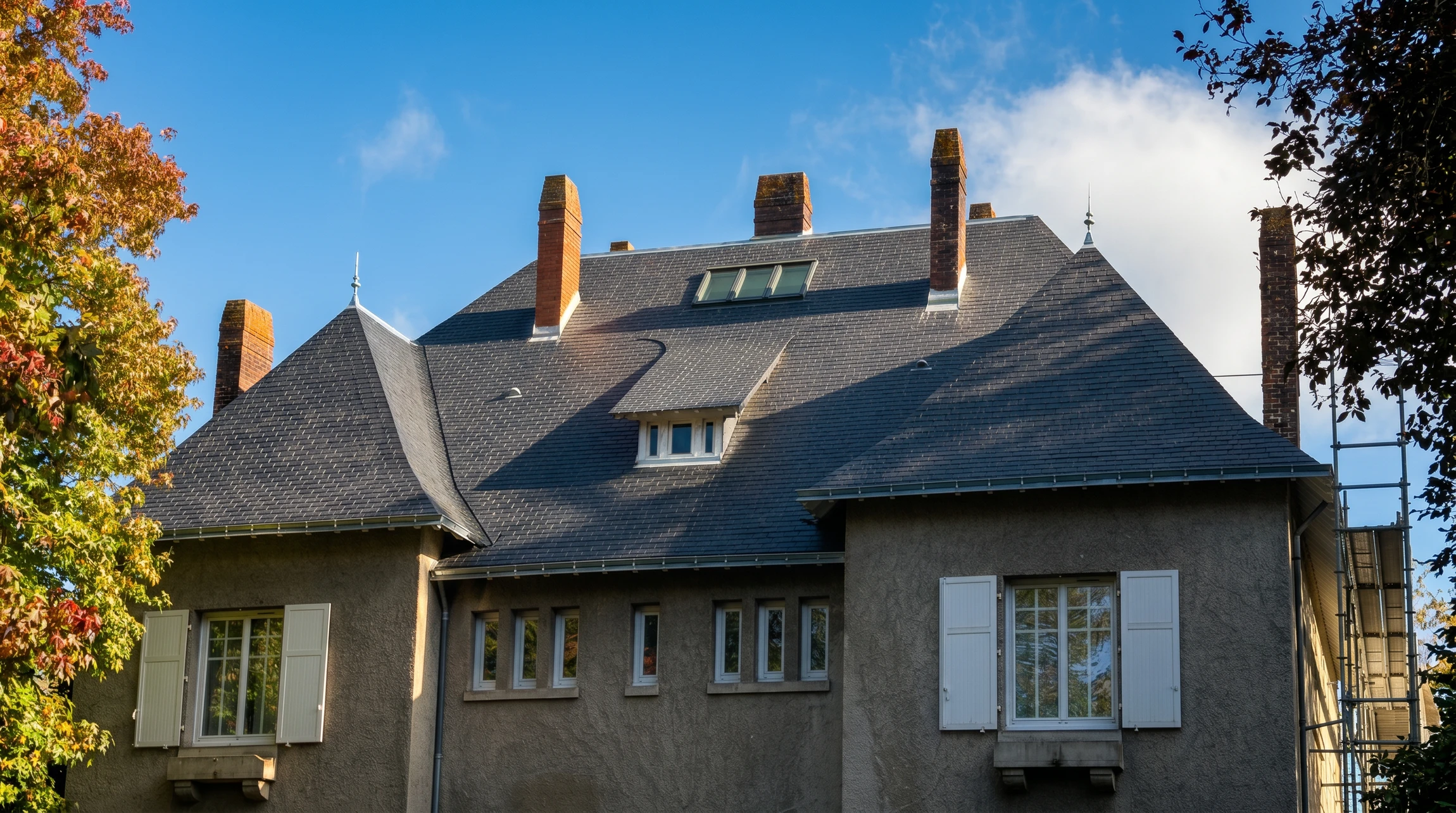 Photographie en perspective d'une toiture en ardoise rénovée, vue en plongée depuis l'extérieur d'une maison résidentielle de type manoir ou demeure traditionnelle. La toiture doit montrer clairement l'ardoise naturelle gris anthracite ou noire avec ses motifs caractéristiques réguliers, accompagnée de détails de charpente ou des ouvrages de couverture. L'image doit capturer le relief et la texture de l'ardoise dans la lumière naturelle, avec un ciel ou un environnement extérieur visible en arrière-plan, illustrant le résultat d'une rénovation de qualité professionnelle.
